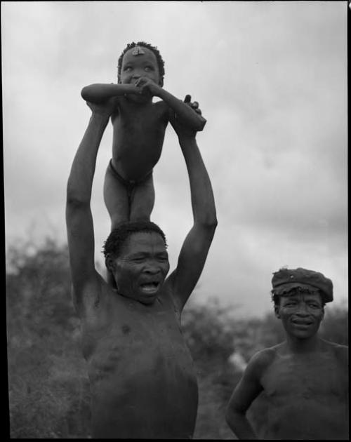 "/Qui Navel" singing medicine songs while holding his son, "Little ≠Gao," on his shoulders and ≠Toma standing to the right wearing a hat