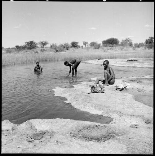 Three men at the edge of a pan, one sitting on the sand, one leaning over with his hand in the water and one bathing