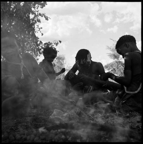 Family sitting next to a fire preparing food, with a child standing next to them