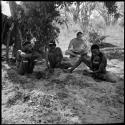 Three men sitting with Nicholas England, one working on arrows, one pounding mangetti nuts, with Nicholas England sitting behind them
