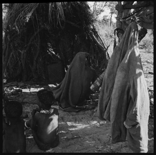 Person covered with a blanket sitting in front of a shelter skerm, with two children sitting behind them, view from behind, Western jacket hanging from a post nearby