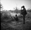 "Old /Gasa" standing, with her grandson ≠Toma (son of Gau) sitting by the fire behind her