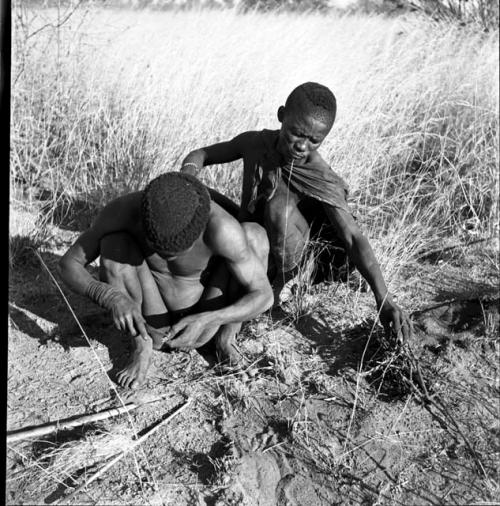 ≠Gao (headman of Band 3) and "/Qui Hunter" starting a fire, with "/Qui Hunter" notching a stick with his knife, and ≠Gao ready to feed twigs to the fire when it starts