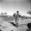 Woman carrying two water bags made from animal stomachs on a digging stick on her shoulder