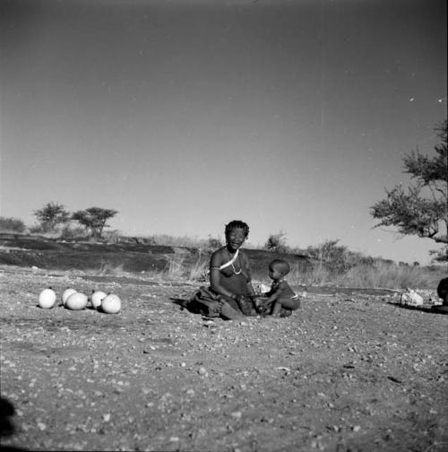 Woman and child sitting near a waterhole, with ostrich egg shells on the ground near them