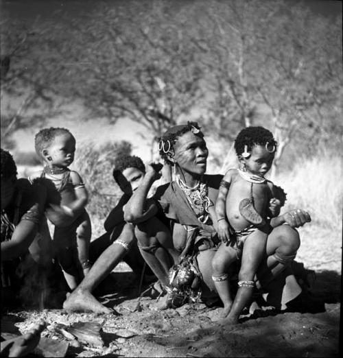 Woman from /Ti!kay's group holding a child on her knee, sitting with a group of women and children