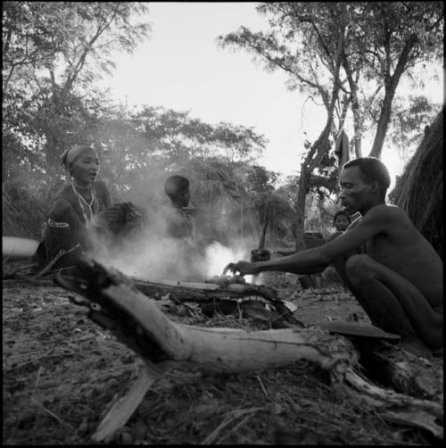 Man cooking mangetti nuts, with a woman and a child sitting across the fire from him