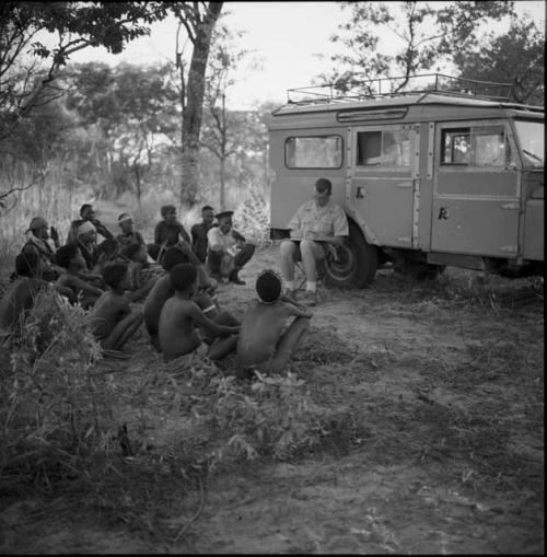 Group of people sitting in a semi-circle around Nicholas England, sitting in front of the expedition Land Rover, taking notes