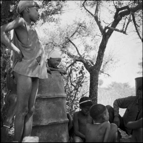 Tsamgao wearing a shirt, standing next to an oil drum, with a group of children sitting next to it