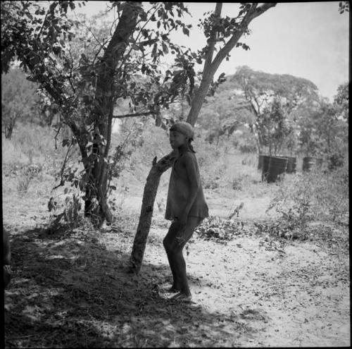 Tsamgao wearing a shirt and a bandana, standing next to a tree, with oil drums in the background