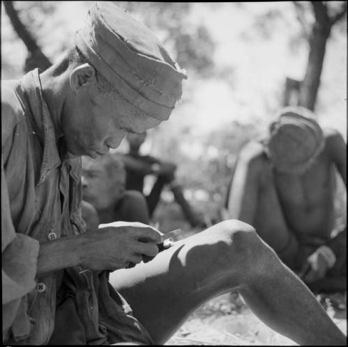 "Short /Qui" sitting, sharpening an arrow point with a metal file, close-up