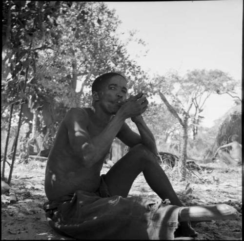 "Short /Qui" sitting, smoking, with a man sitting in front of a skerm in the background