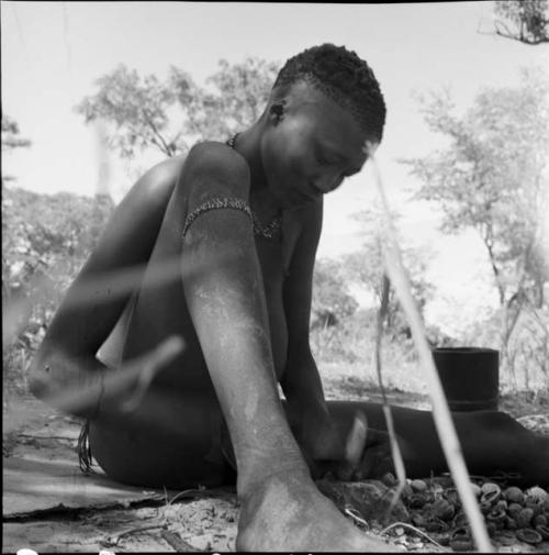 Man wearing a bracelet on his leg, sitting, cracking mangetti nuts on a stone, with the nutshells on the ground in front of him