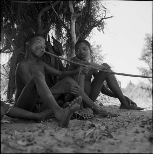 Boy playing a musical bow, sitting with another boy in front of a skerm constructed for hot weather