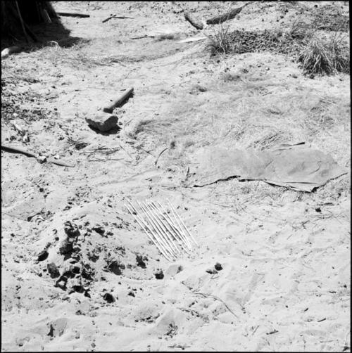 Arrows lying next to an animal hide on grass and sand, with a fire paddle, arrows and two logs in the background