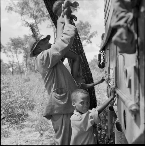 /Ti!kay lifting a child to see inside an expedition truck, with a boy standing next to him looking toward the camera