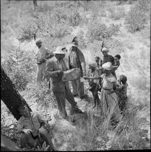 Group of people standing around an expedition member who is pouring water from a canvas bag into a cup held by a girl, with Nicholas England sitting against a tree near them