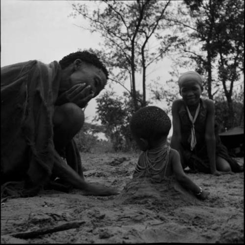 N!ai and an unidentified woman play with a baby sitting in a hole in the sand, view from behind