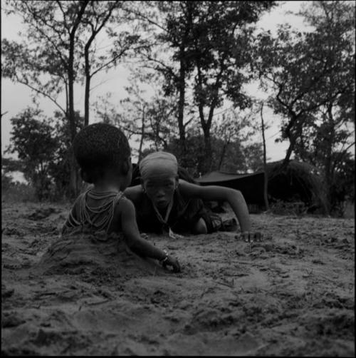 N!ai playing with a baby sitting in a hole in the sand, with an expedition tent and a skerm in the background