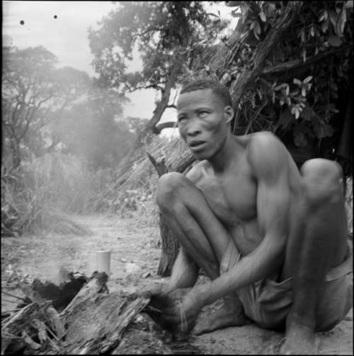 Man holding a piece of wood in his hands, squatting in front of a fire