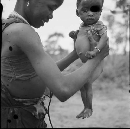 N!ai standing, holding a baby who has just urinated, with wet feet and legs