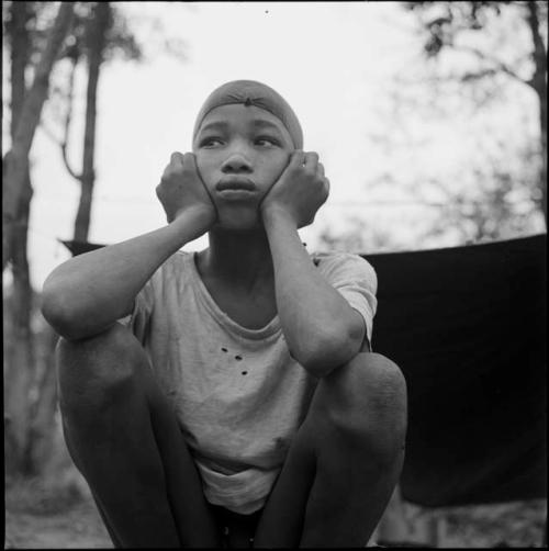 Tsamgao wearing a T-shirt and a bandana, squatting, holding his face in his hands