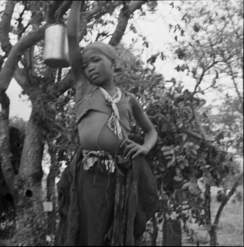 N!ai wearing a kaross and a halter top, standing next to a tree with a tin pail hanging in it