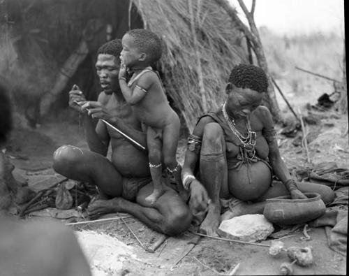 "Gao Medicine" binding an arrow shaft with sinew, with /Gaishay leaning on him, and Di!ai sitting next to him pounding something with a stone