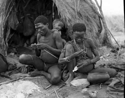 "Gao Medicine" binding an arrow shaft with sinew, with /Gaishay leaning on him, and Di!ai sitting next to him pounding something with a stone