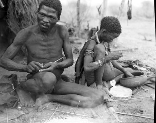 "Gao Medicine" binding an arrow shaft with sinew, with Di!ai sitting next to him pounding something with a stone