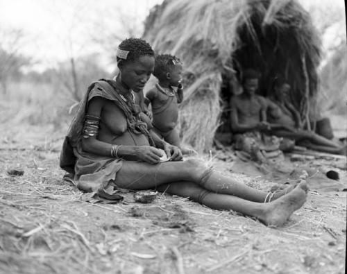 Woman sitting in front of Gau's skerm, with a child standing next to her, Gau and one of his wives sitting in the skerm