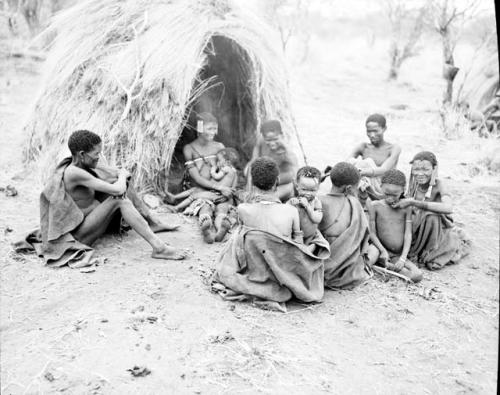 "/Qui Hunter" of Band 2 sitting in front of a skerm with his wife, /Naoka, holding her baby girl, /Khoa, and a group of people including Dam (husband of ≠Gisa, /Naoka's sister), Be (wife of Gau) with her son, and "Gao Scarf"