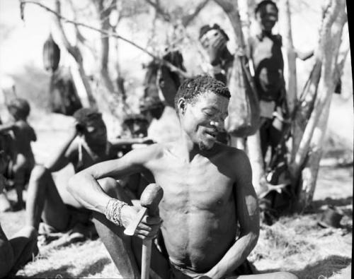 /Ti!kay sitting, holding an axe, with a group of people behind him