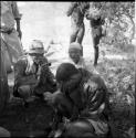 /Ti!kay wearing Western clothing, holding a pipe in his mouth, squats next to Venter who is working on tobacco