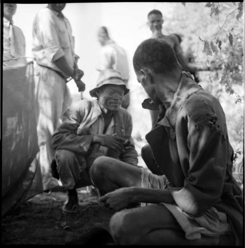 /Ti!kay wearing Western clothing, holding a pipe in his mouth, squats next to Venter who is working on tobacco