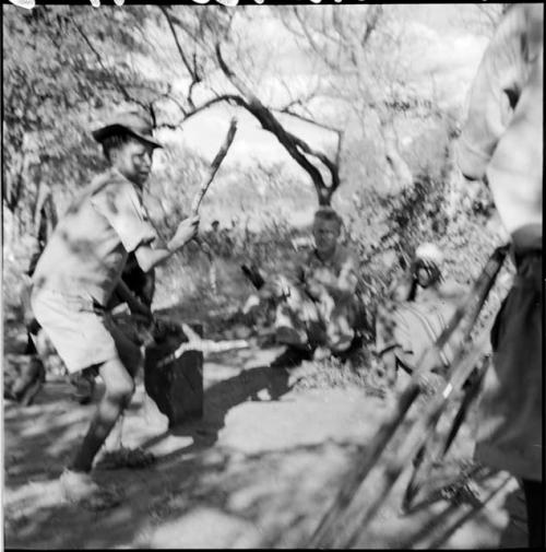 Tsamgao wearing Western clothing, playing with a stick, with John Marshall filming him, two people sitting in the background watching