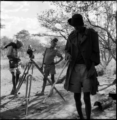 Venter standing next to a tripod with a film camera, holding a stick, with Tsamgao and /Gaishay wearing Western clothing, standing in the background