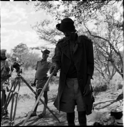 Venter standing next to a tripod with a film camera, holding a stick, with Tsamgao and /Gaishay wearing Western clothing, standing in the background