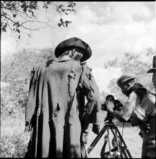 Venter wearing a tattered coat, standing, view from behind, with /Gaishay focusing the film camera in the background