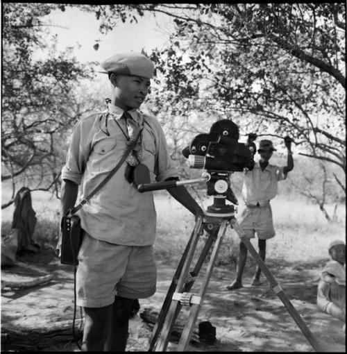 /Gaishay wearing Western clothing, standing next to a film camera on a tripod, with a man standing in the background