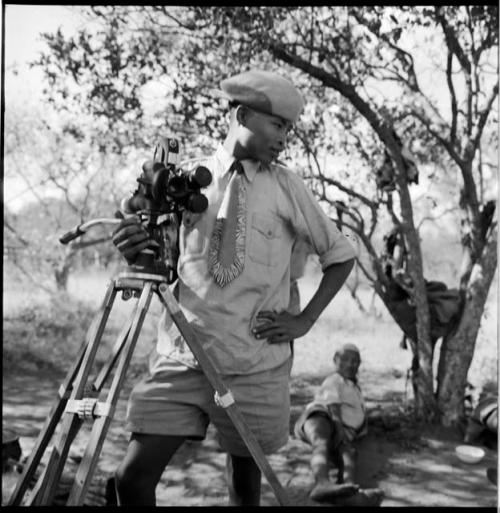 /Gaishay wearing Western clothing, standing next to a film camera on a tripod, with a person sitting in the background