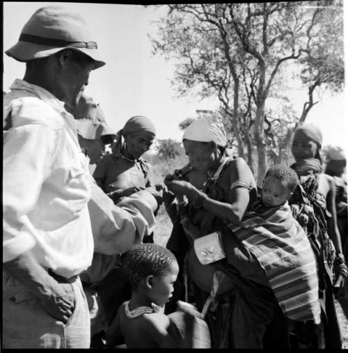 Woman with a baby tied to her back with a striped cloth, lighting a pipe, standing with a group of people, including /Ti!kay