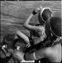 N!ai adjusting her headband, sitting with another woman at the edge of a pan, view from behind