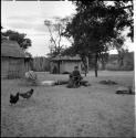 Child sitting with a staff member by a fire, with chickens near them, skerms in the background