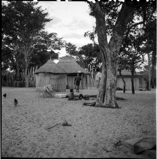 Child sitting at a fire, with two expedition members standing nearby, chickens, skerms in the background