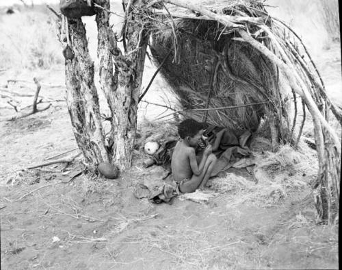 Boy sitting under a skerm built against a tree for shade