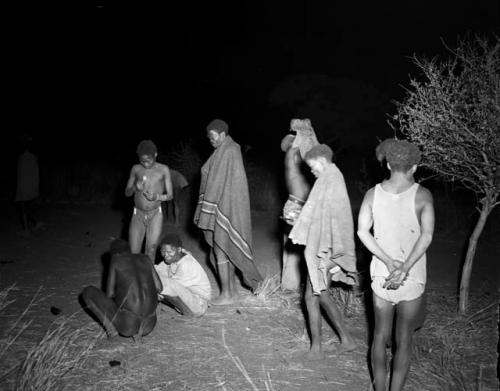 Group of people in an intermission in a night dance, including "Gao Medicine" wrapped in a blanket, /Gunda (N!ai's husband) standing in front of him, //Ao wearing a white shirt and ≠Toma wearing a white shirt and shorts, with his back to the camera