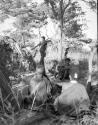 Group of people sitting, with a man carrying wood behind them