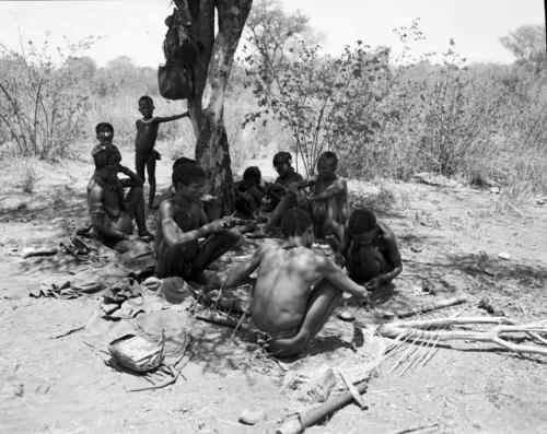 "/Qui Navel" and ≠Gao working on arrows at the werft of ≠Gao (headman of Band 3, son of /Gasa and brother of Gau), sitting with /Goishay (≠Gao's wife), "/Gao Music" (brother of /Goishay) playing the //guashi, ≠Toma (≠Gao and /Goishay's son), Dam (husband of ≠Gisa), Khwo//o (mother of "/Gao Music" and /Goisha) and /Naoka (wife of "/Gao Medicine")