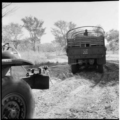 Man sitting on top of an expedition truck, with the truck pulling another truck with a rope, view from behind